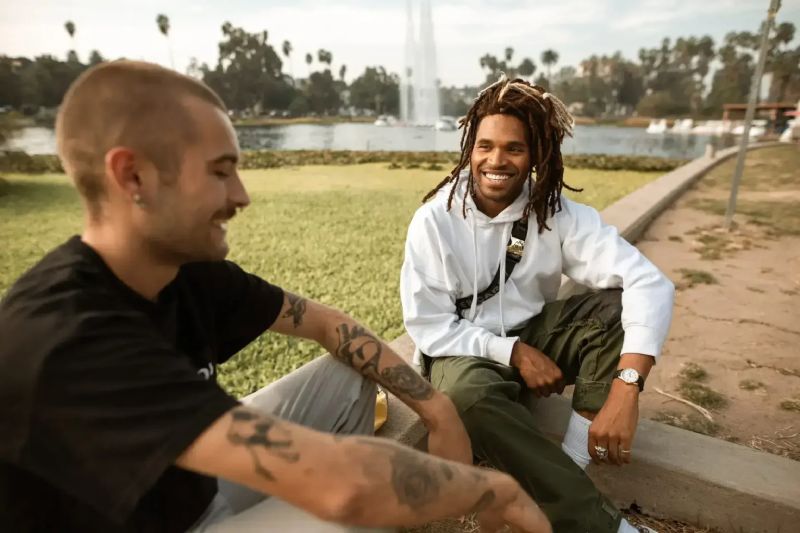 Two men casually talking by a waterfront park, smiling and enjoying their conversation, with greenery and a fountain in the background, reflecting a relaxed social setting.