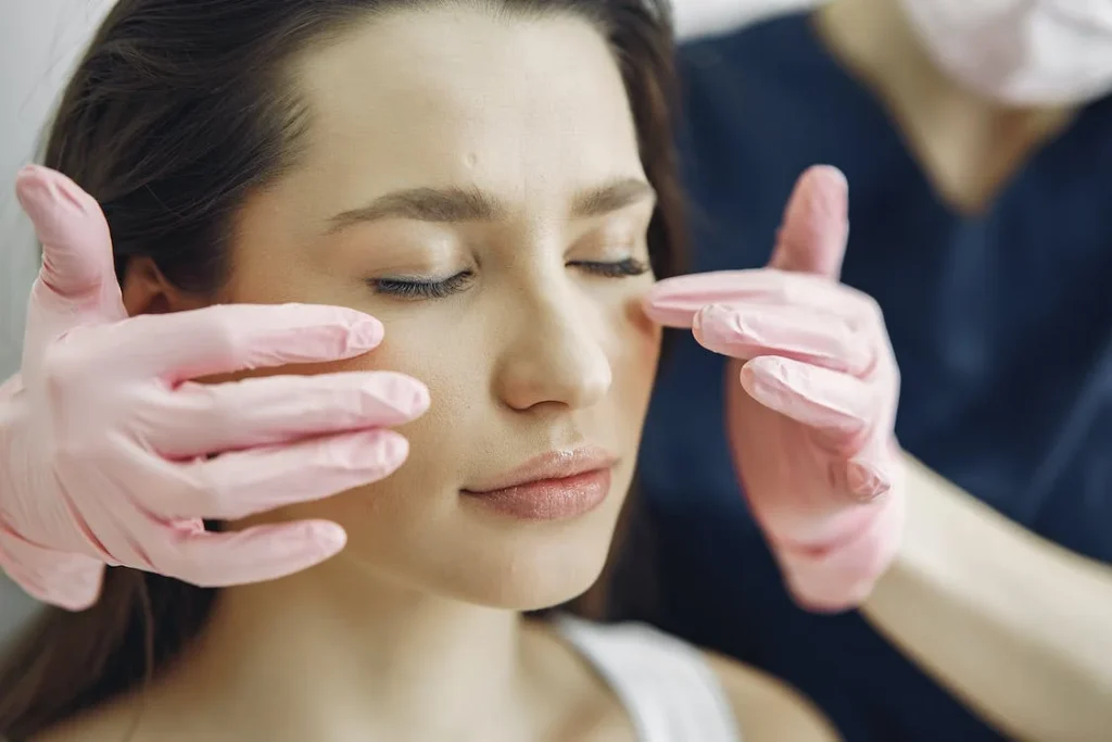 Woman receiving facial treatment from a skincare professional, hands with pink gloves gently assessing skin, highlighting post-Lumecca IPL care and recovery.