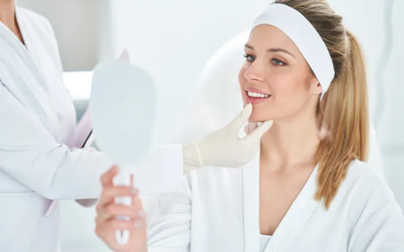 Woman in a spa setting smiling at her reflection in a mirror while a professional applies Botox treatment, showcasing a relaxed and refreshed appearance for holiday beauty preparation.
