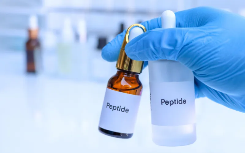 Hand holding two peptide therapy vials, one amber and one clear, labeled "Peptide," in a clinical setting related to HGH peptide therapy for anti-aging.