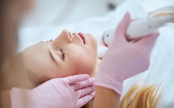 Woman receiving microneedling treatment on her face, with a practitioner using a microneedling device, both wearing gloves, in a skincare clinic setting.