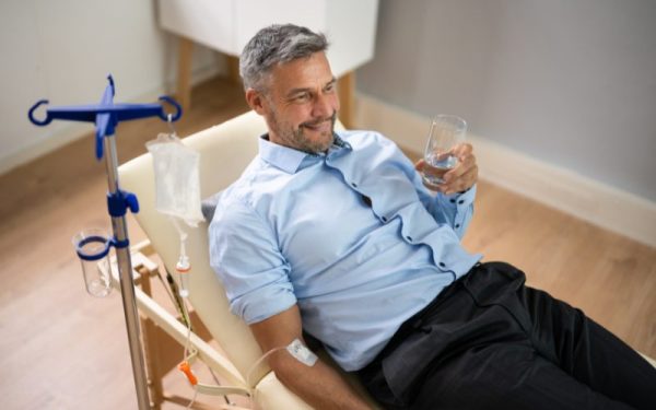 Man receiving IV ketamine therapy, smiling in a chair while holding a glass of water for PTSD treatment relief